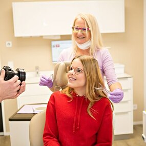 Junge Frau beim Zahnarzt lächelt, während Zahnarzthelferin und Fotograf anwesend sind.